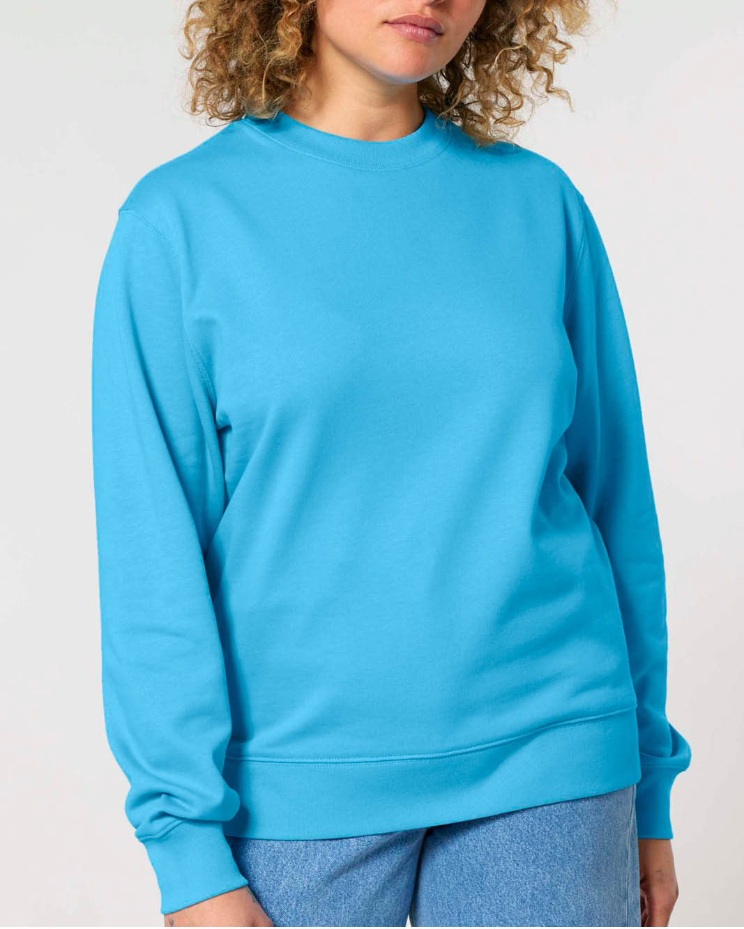 A woman wearing a bright turquoise blue crewneck sweatshirt, showcasing it's oversized fit, cuffed sleeves, waistband and neckline.