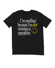 A gender-neutral black running crewneck t-shirt with a graphic text print on the front reading 'I'm smiling because I'm not running a marathon.' in white and yellow, accompanied by a yellow smiley face and the Raw Threads X Smiley logo.