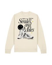A natural sweatshirt featuring a graphic print on the front with the text 'Smiles and Miles', a black retro smiley face, a white sneaker illustration, and small yellow Smileys. Features the text: self-love, good runs ahead.