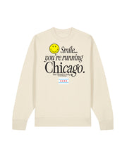 A natural tan gender-neutral crewneck sweatshirt featuring a graphic print on the front with a yellow smiley face and the text 'Smile you're running Chicago' including the Chicago flag and the Raw Threads x Smiley logo.