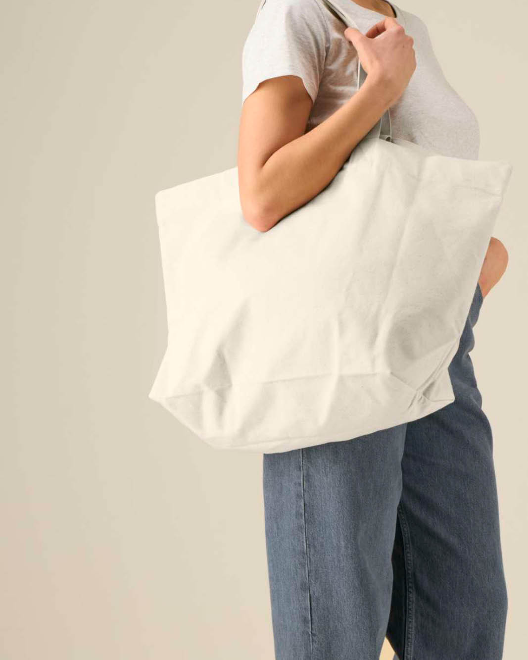 A person carrying a large natural canvas tote bag over their shoulder to show how large the canvas bag is, and that you can use the shoulder handles.
